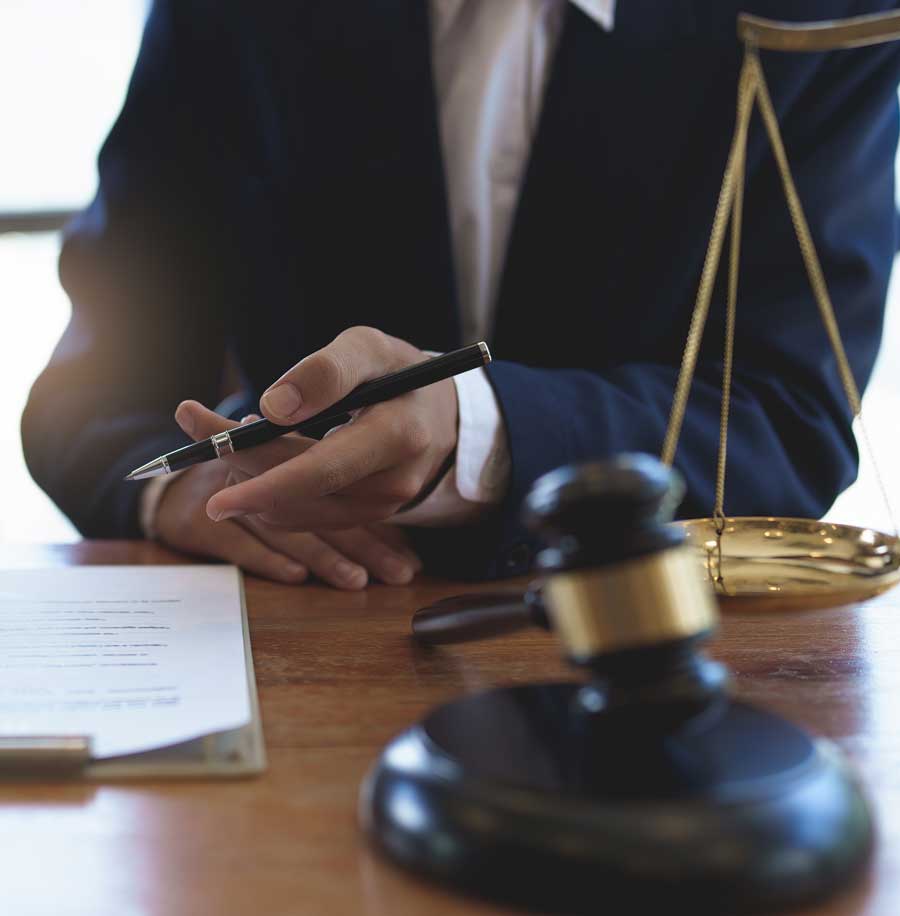 A man in a dark blue suit holding a pen over a table with a judge's gavel and set of golden scales
